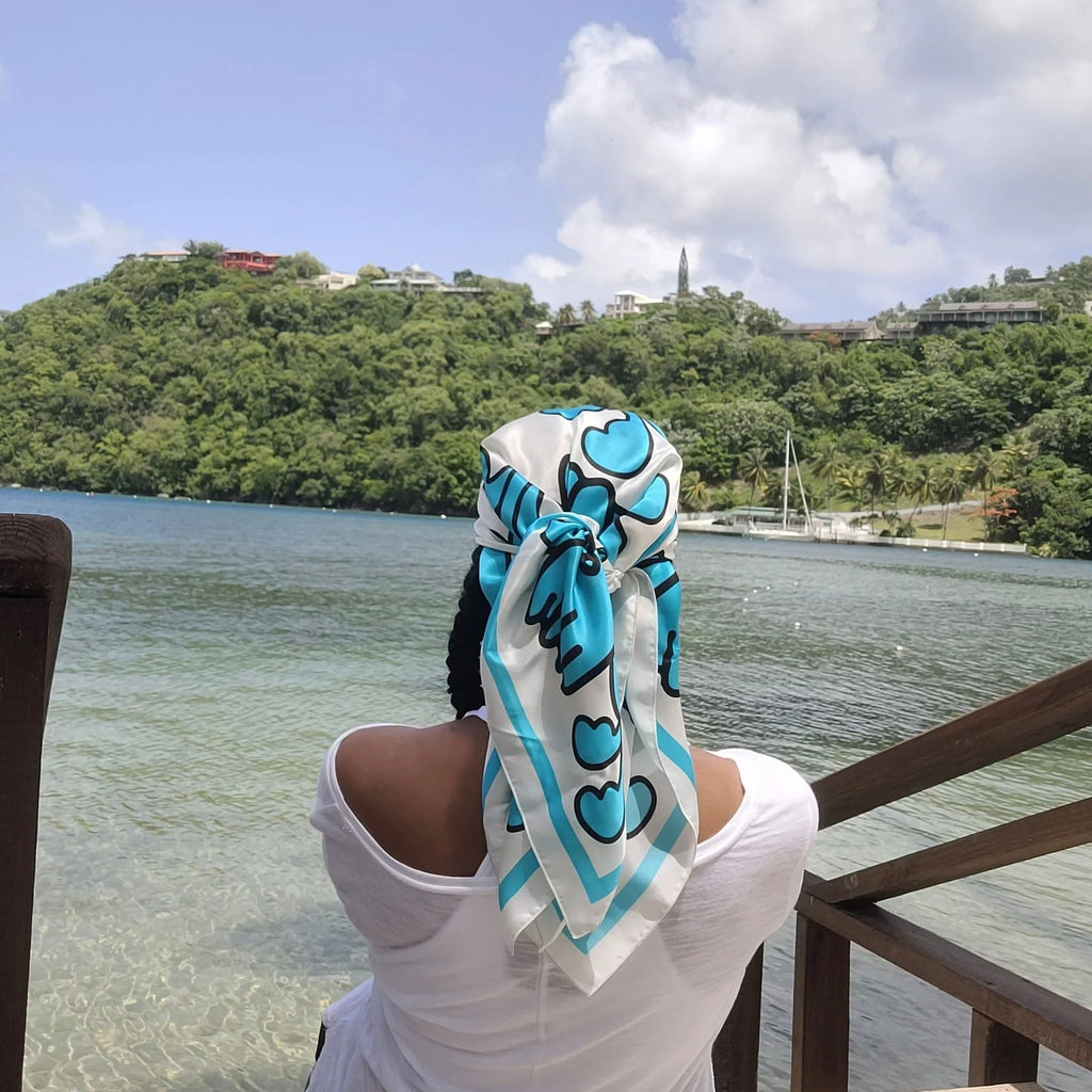 A woman wearing a Caribbaby silk scarf wrapped around her head sitting on a beach looking at the water.